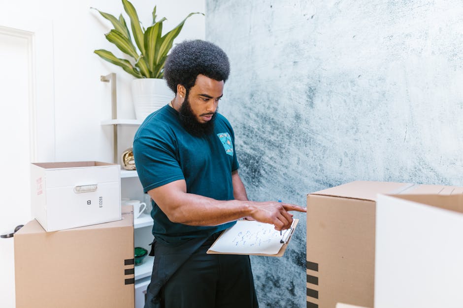 A man with a dark curly afro hairstyle, wearing a dark teal T-shirt, is standing inside a room with textured light blue walls, surrounded by unpacked cardboard boxes of various sizes for house or office packing. He is holding a clipboard with documents or a moving checklist in one hand and pointing to the paper with his other hand, indicating a discussion or review of the packing process. Behind him, there is a white shelving unit with a large potted plant with long green leaves, a white ceramic mug, and a small decorative item. The boxes are positioned on the floor and some are stacked, suggesting the ongoing process of packing or organizing items for a house relocation. The environment appears well-lit with natural light, and the scene reflects a professional yet straightforward approach to packing and moving logistics, as offered by Man and Van Camberwell’s removals service in Southwark.