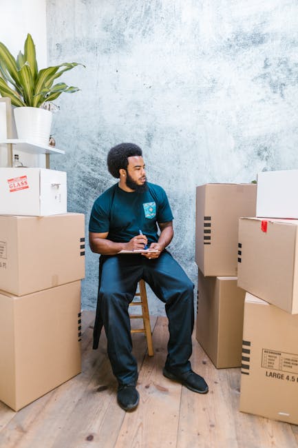 A man wearing a yellow T-shirt and blue jeans is standing in a room during a home relocation process, holding a clipboard with a pen and making notes. Behind him, there are several cardboard moving boxes, some open and some sealed, arranged on a light wooden floor. To his left, there is a tall potted plant with green leaves positioned against a white wall. The scene appears to be inside a residential property, with the boxes ready for packing or unpacking, indicating the early stages of furniture transport and moving logistics facilitated by Man and Van Camberwell. The image captures the packing and moving process in a professional and neutral environment, emphasizing careful planning and organization crucial to house removals and business relocations.