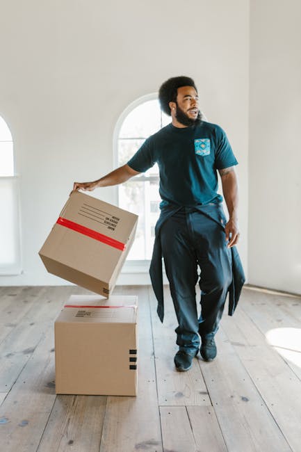 A man with dark curly hair and a beard, dressed in a dark blue T-shirt and matching trousers, stands in a bright, empty room with white walls and large arched windows. He is holding a cardboard box wrapped with red packing tape in his right hand, preparing for a home relocation. In front of him are two additional cardboard boxes, one stacked on top of the other, both sealed and ready for transport. The room features light wooden flooring and natural light streaming through the windows, creating a clean, minimal setting typical of a house being prepared for moving. The presence of multiple boxes and the man's posture suggest he is engaged in packing or unpacking as part of furniture transport or overall packing and moving activities, with a focus on careful handling to ensure the safety of household items. This scene illustrates a professional or personal packing process associated with house removals, which is a service offered by Man and Van Camberwell, supporting the page about costs and quotes for house removals in Camberwell SE5.
