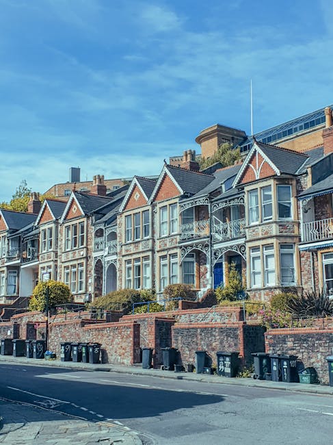 A row of Victorian-style terraced houses with brick facades, white decorative trim, and bay windows, situated on a residential street in Camberwell. In the foreground, there is a paved pavement with black and blue wheelie bins lined along the curb. The houses are positioned behind a low brick wall, with small landscaped front gardens featuring bushes and greenery. The sky above is clear and blue, indicating a bright day, and the image depicts an urban area typical of home relocation environments, where interior furniture and moving boxes might be transported during house removals by companies such as Man and Van Camberwell. The scene captures the exterior setting before or after a moving process, with no visible activity but suggesting a calm residential street ready for a furniture transport or packing and moving operation.