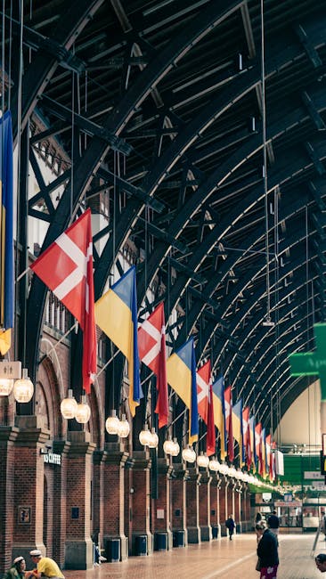 The interior of Denmark Hill station with a high, arched ceiling made of metal beams painted dark blue, adorned with multiple flags in red, yellow, blue, and white hanging vertically along the platform. The platform features brick walls with vintage-style light fixtures and digital display boards. In the foreground, a person wearing dark clothing and a hat is walking near the edge of the platform, while in the background, another individual is seen carrying a box. According to the structure and environment, this setting highlights the loading and unloading process associated with home relocation or moving services. For a professional move, Man and Van Camberwell would manage the packing, loading, and transport of furniture, boxes, and appliances, utilizing suitable equipment such as trolleys, straps, and blankets to ensure safe and efficient transportation. This scene emphasizes the logistical aspects of house removals, particularly the movement of items through station platforms during a furniture transport or packing and moving operation.
