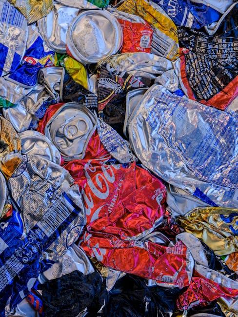 Close-up view of a pile of crumpled aluminium soda cans and crushed beverage packaging made of plastic and aluminium, positioned on pavement outside a property. The collection includes various soda brands with identifiable branding such as Coca-Cola and other snack wrappers, some with pull-tabs visible. The cans are in different states of compression, with some partially flattened, while others maintain a more rounded shape. The background suggests a disposal or recycling area used during a home relocation or removal process, with cans stacked tightly together, indicating recent collection for recycling. This scene reflects the waste disposal stage following a house move, with Man and Van Camberwell facilitating the removal process including e-waste, packaging materials, and recyclables, supporting sustainable disposal and reuse practices in Camberwell.