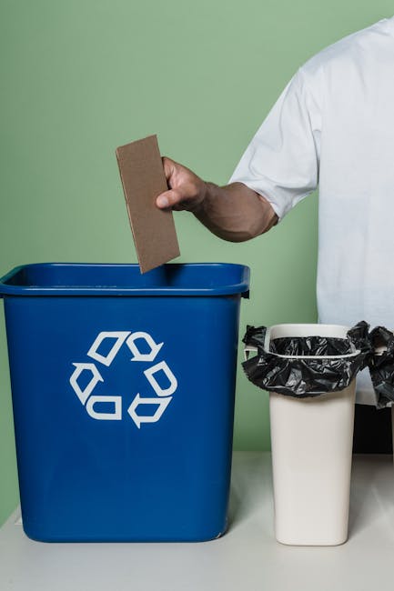 A person wearing a white shirt holding a small piece of cardboard above a blue recycling bin with a white recycling symbol on the front, positioned on a table against a light green wall. Next to the blue bin is a white trash can lined with a black plastic bag, suggesting the process of waste separation and disposal following a home relocation or furniture transport. The scene highlights proper sorting of packaging materials, cardboard, and general waste during the decluttering or packing phase of house removals, as managed by Man and Van Camberwell. The lighting is even, illuminating the objects clearly, and the environment appears clean and organized, emphasizing responsible recycling and waste management during the moving process.