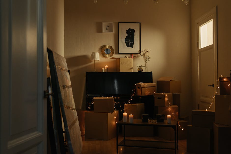 Inside a room prepared for a home relocation, with a black upright piano positioned near the wall, surrounded by multiple cardboard boxes of various sizes, some stacked on top of each other and others placed on the floor, indicating packing for a furniture transport process. The boxes are partly wrapped in plastic and sealed with packing tape. An open doorway in the foreground frames the scene. The room features warm ambient lighting from candles and a small table lamp, creating a soft glow, and decorative elements such as a framed artwork depicting a stylized human figure, a round mirror with a sunburst frame, and a tall plant with thin branches in a vase on top of the piano. The window on the right allows natural light into the space, which is organized for a smooth loading process by [COMPANY_NAME], showcasing their expertise in house removals and professional packing and moving services from Camberwell.