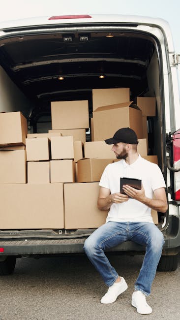 A man wearing a black cap, white t-shirt, and blue jeans is sitting on the pavement next to the open rear doors of a moving van. Inside the van, there are multiple cardboard boxes of various sizes, some stacked on top of each other, with a few secured by packing materials such as plastic wrap or paper. The boxes are arranged along the interior walls of the van, ready for a home relocation or furniture transport process. The scene takes place outdoors, with natural light illuminating the area. The man is holding a tablet, possibly coordinating loading or packing tasks as part of a house removal service. This image illustrates the loading process involved in professional removals, supported by a company like Man and Van Camberwell, which specialises in removals and moving logistics.