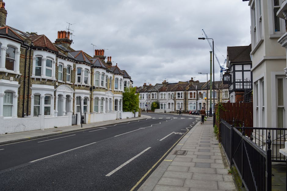Photograph of a residential street in Camberwell showing a row of terraced Victorian-style houses with bay windows and chimneys under an overcast sky. The street features a paved sidewalk with a black metal railing fence on the right and a cyclist and pedestrians walking along the pavement. The road is marked with white dashed lines, and several parked cars are visible further down the street. In the background, there are construction cranes indicating ongoing building work, and streetlights line the sidewalk. This scene captures an urban environment typical of a house relocation or moving process, with no vehicles or furniture currently visible. Man and Van Camberwell, a professional removals service, operates in this area and may facilitate residential moves involving furniture transport, packing, and loading procedures in such neighbourhoods.