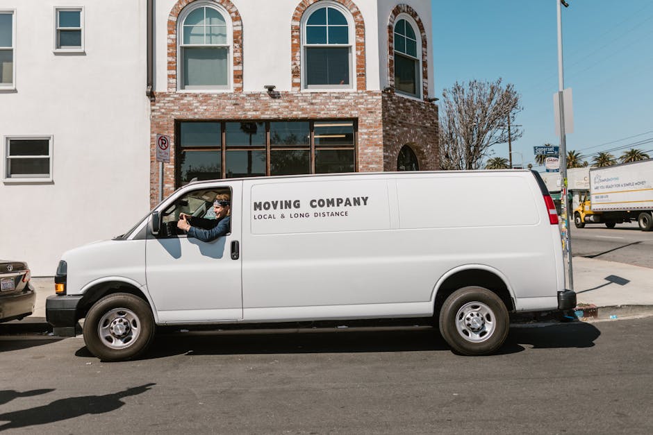 A white moving van with the text 'MOVING COMPANY LOCAL & LONG DISTANCE' printed on its side is parked along the street in front of a brick building with large windows and arched window frames. The dri